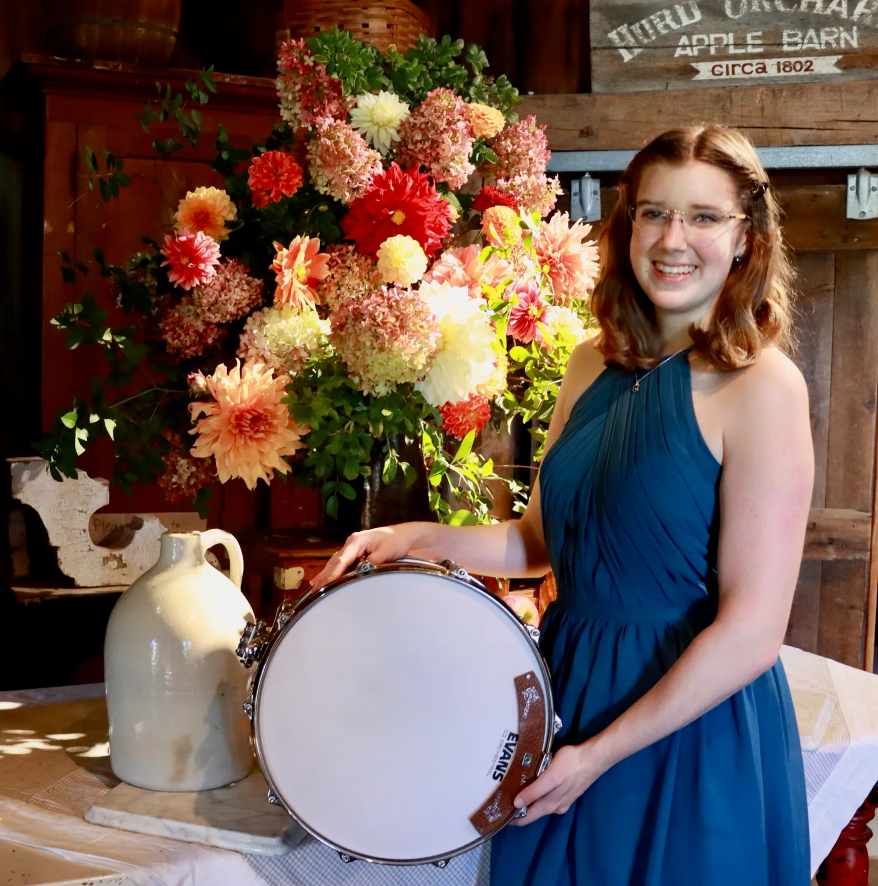 Liz Morad smiling beside a snare drum and a large flower arrangement, wearing a teal-blue dress